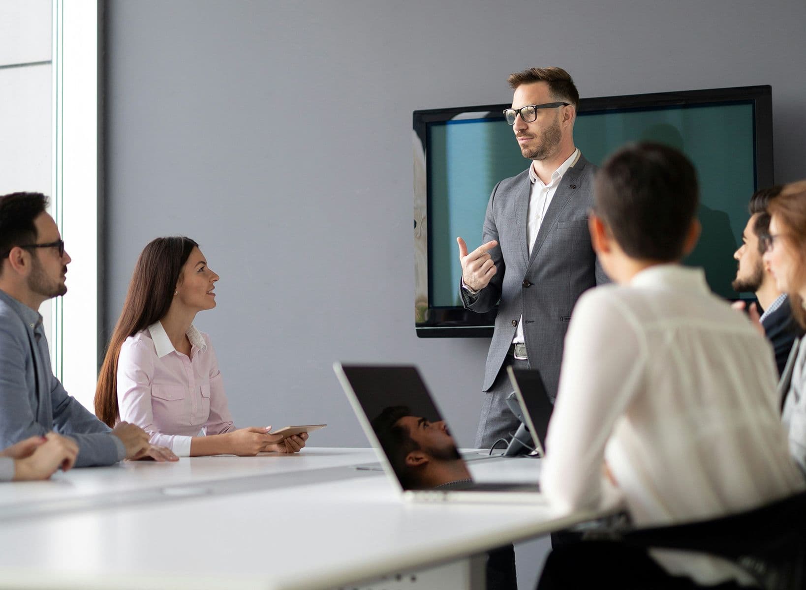 Homme en costume donnant une présentation devant un écran à un groupe de collègues assis autour d’une table.
