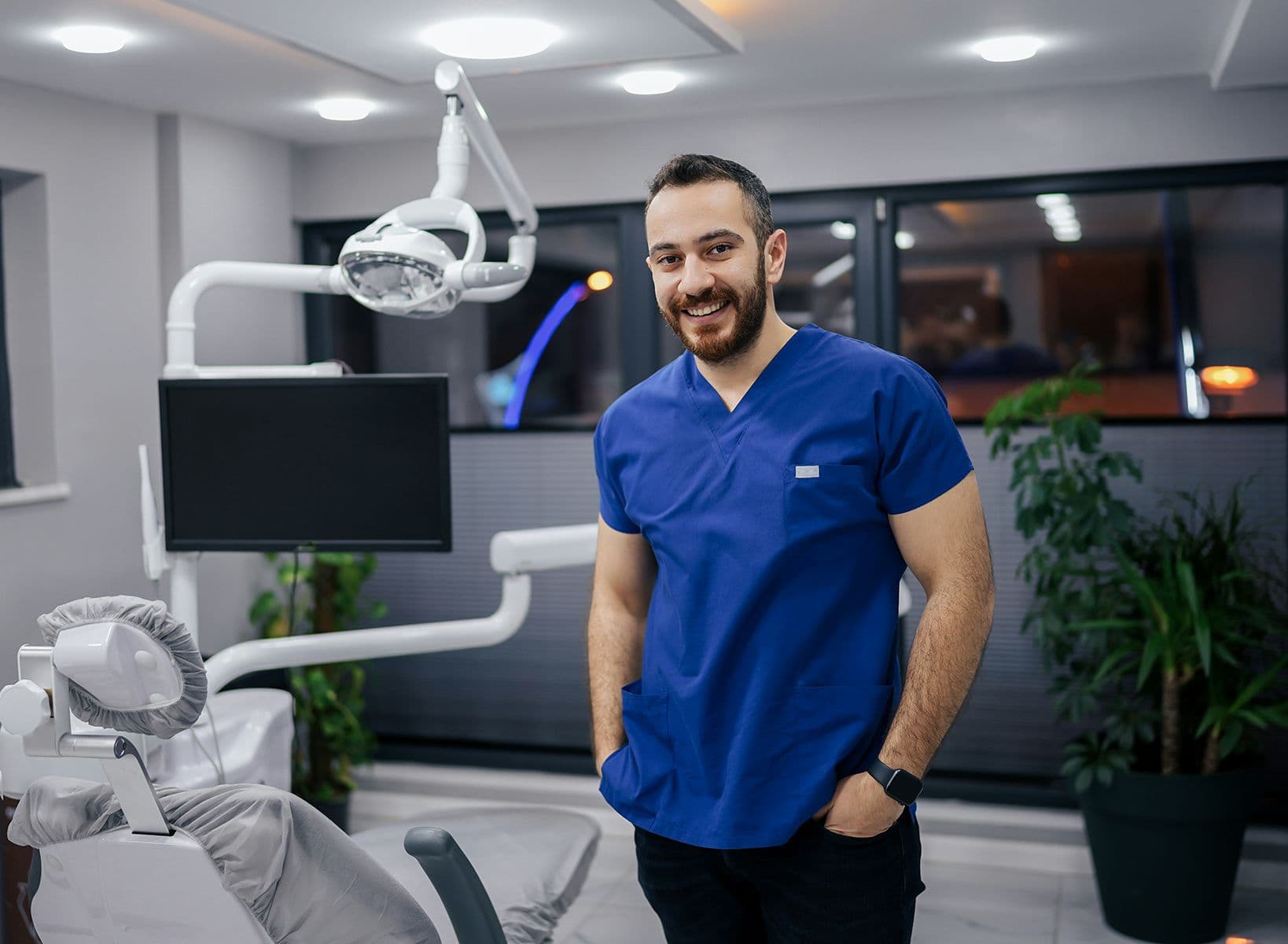 Dentiste souriant en tunique bleue debout dans un cabinet moderne, à côté d’un fauteuil dentaire et d’équipements médicaux.