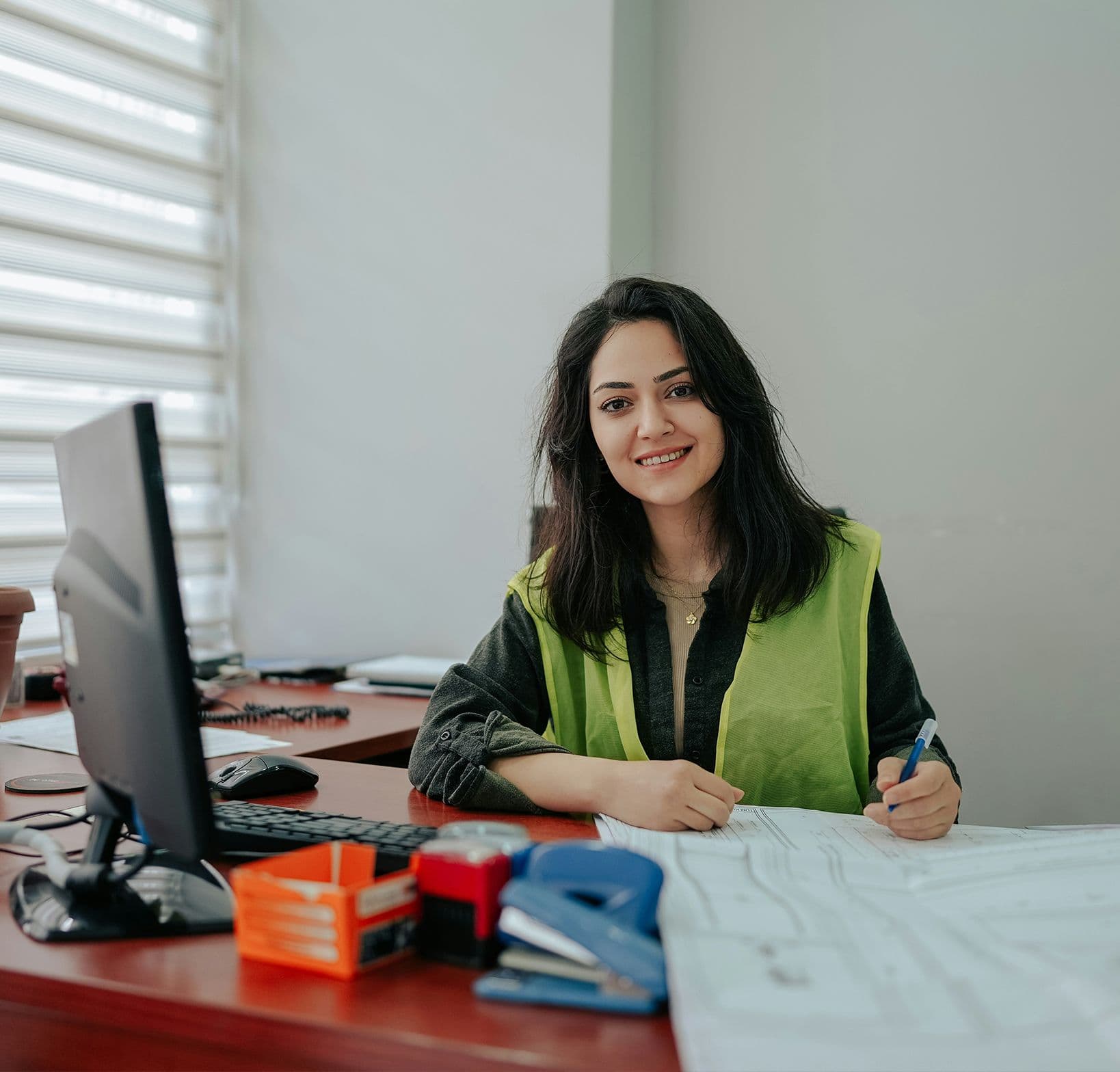 Femme souriante en gilet de sécurité assise à un bureau avec plans et ordinateur, dans un bureau lumineux.
