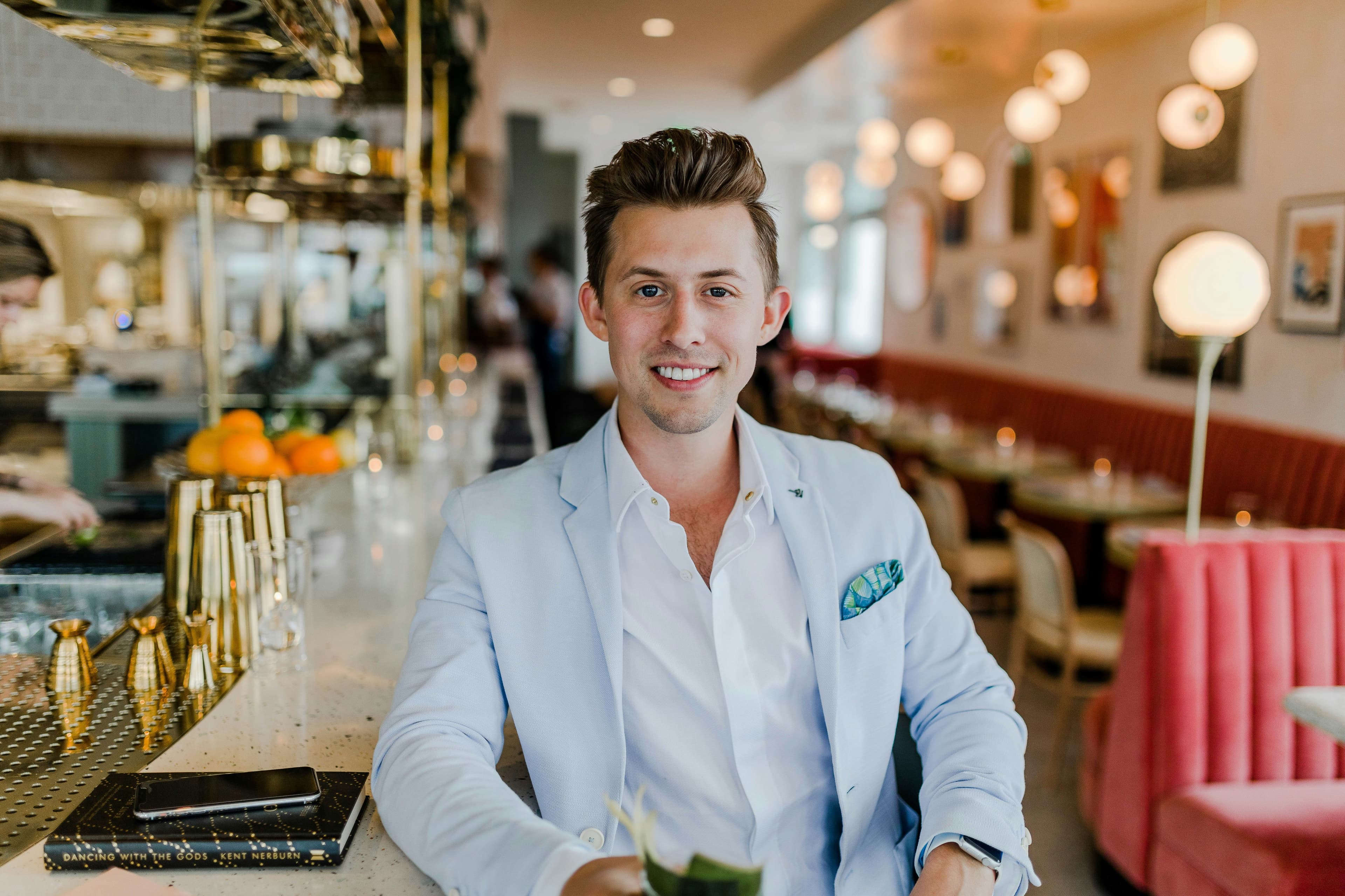 Homme souriant en blazer clair assis au bar d’un restaurant chic, avec une ambiance chaleureuse en arrière-plan.