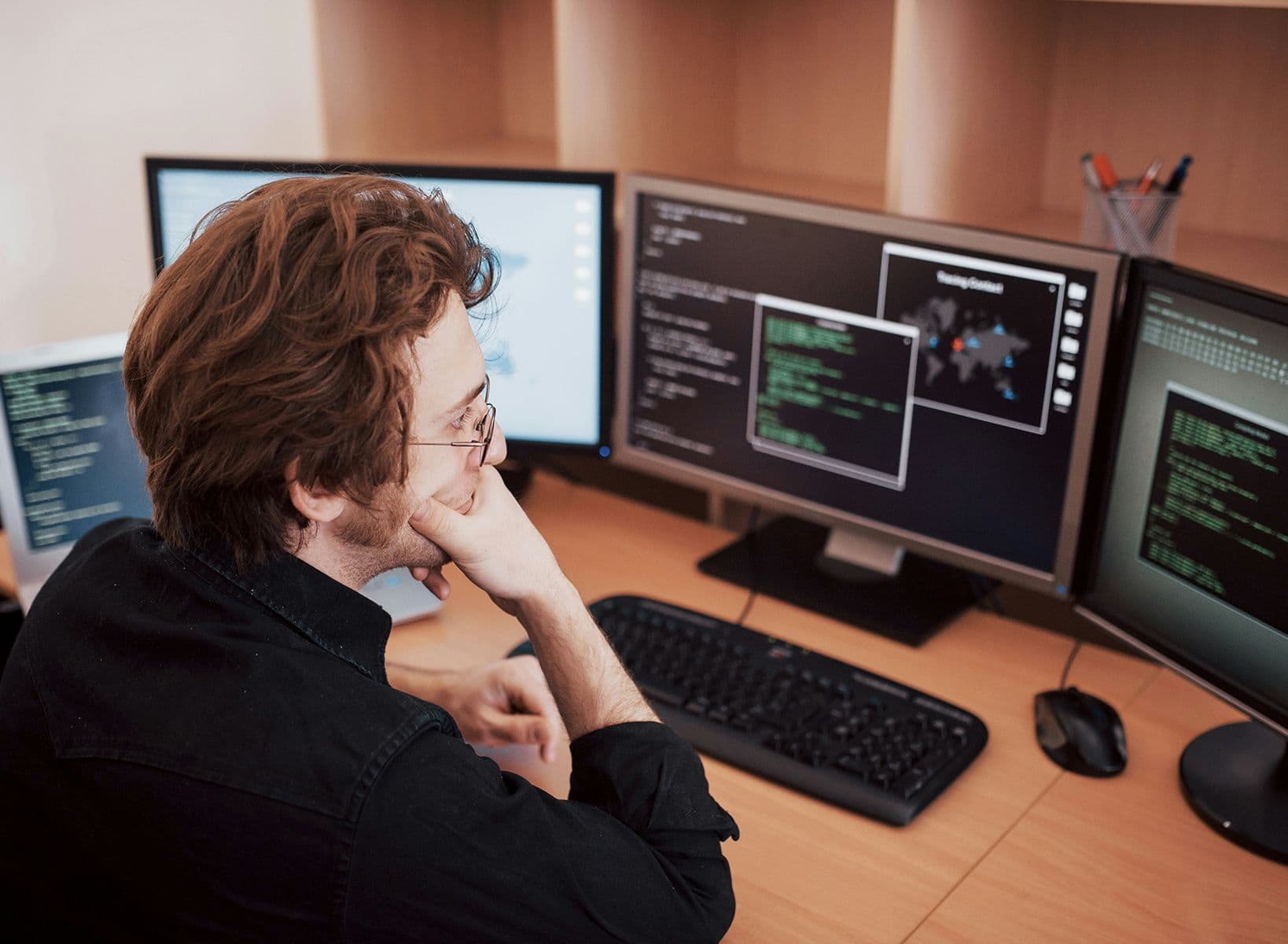 Homme concentré devant plusieurs écrans affichant du code et des données, assis à un bureau informatique.
