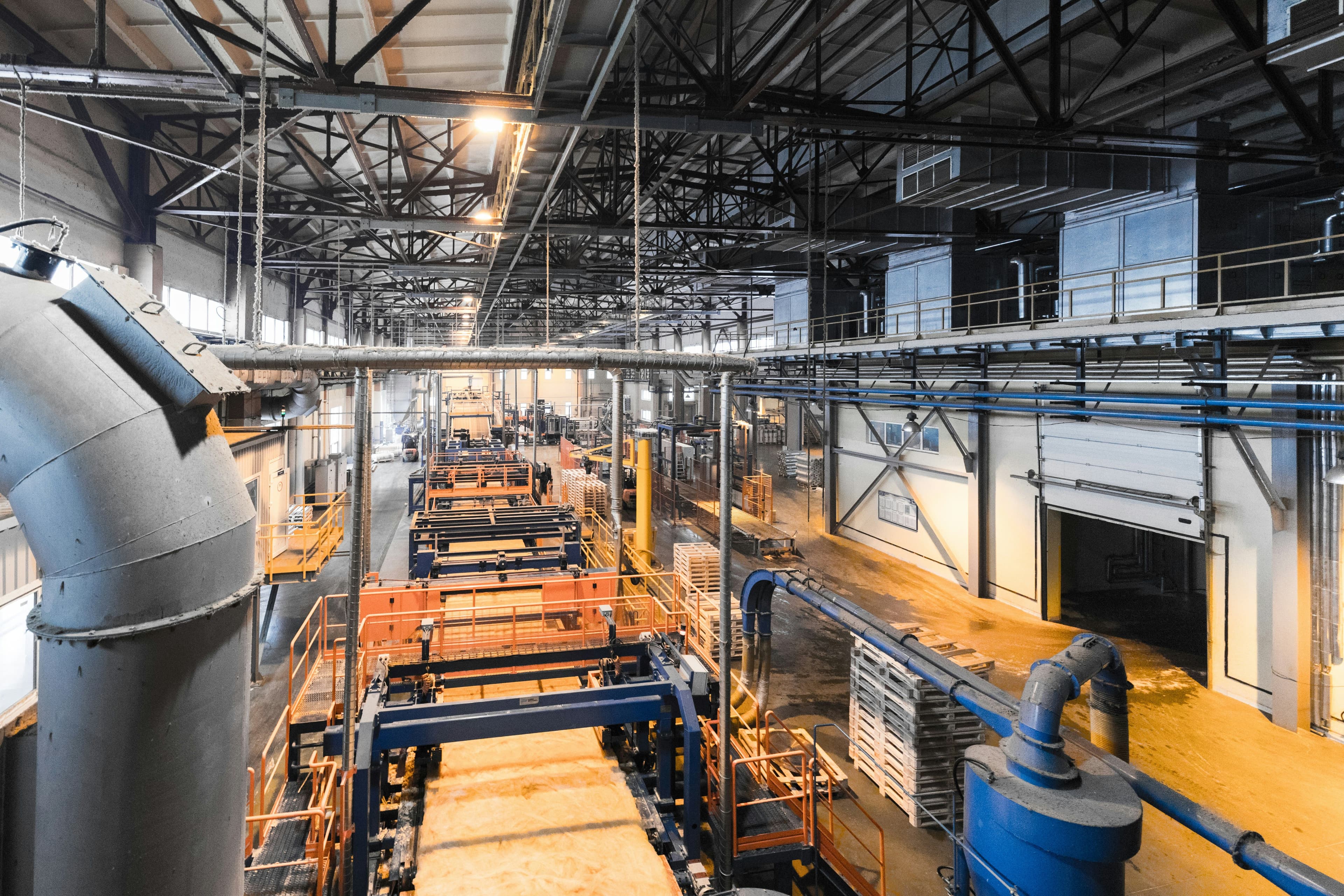 Intérieur d’une usine industrielle avec machines de production, tuyauterie apparente et structure métallique au plafond.
