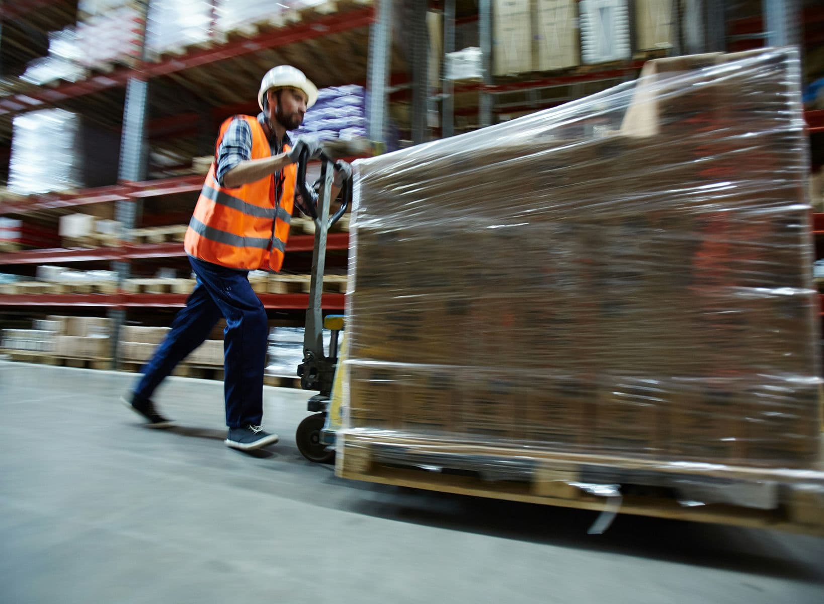 Magasinier en gilet orange et casque poussant un transpalette chargé de cartons filmés dans un entrepôt logistique.