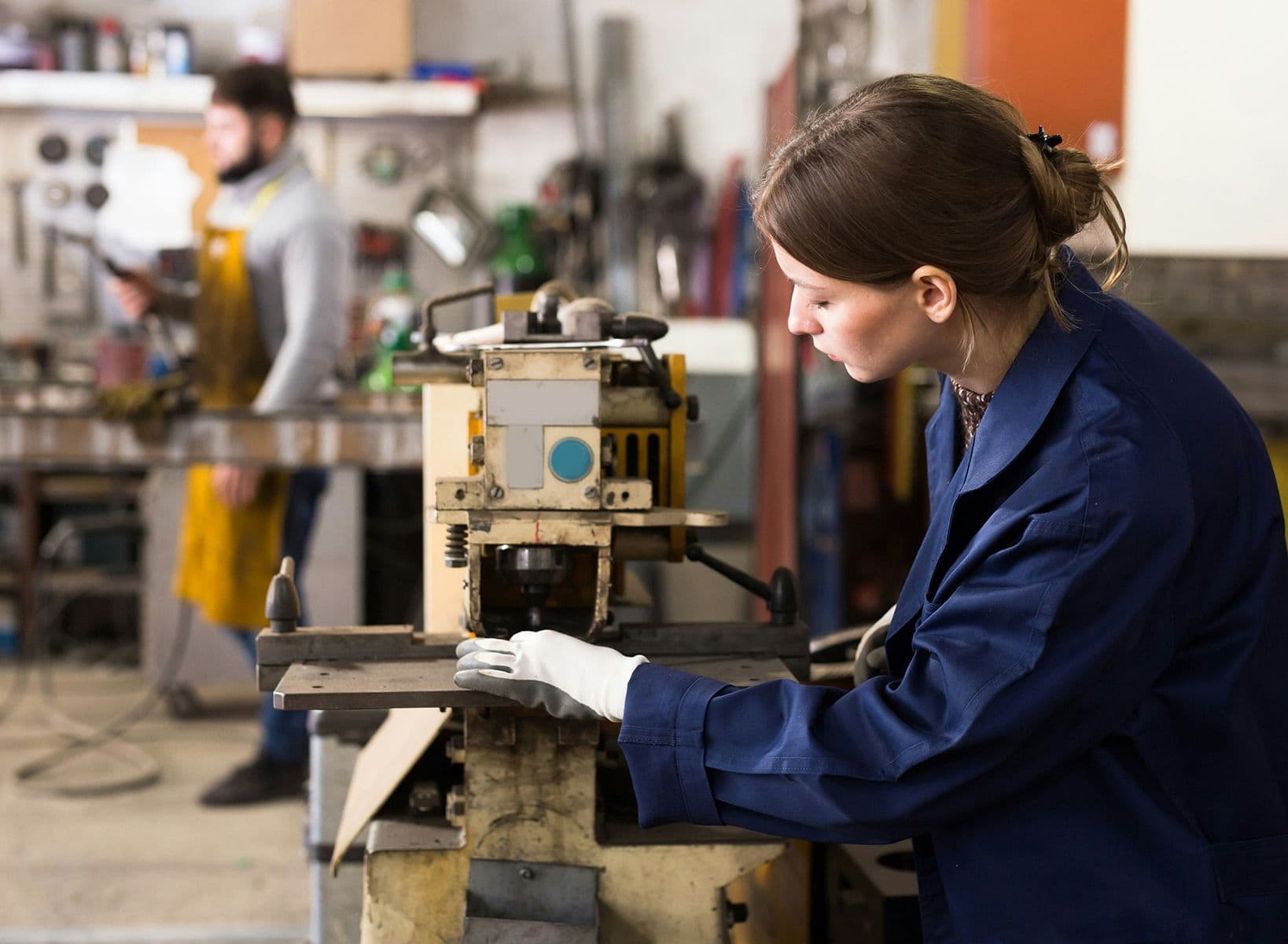 Femme en blouse bleue utilisant une machine-outil dans un atelier, concentrée sur une pièce métallique à usiner.