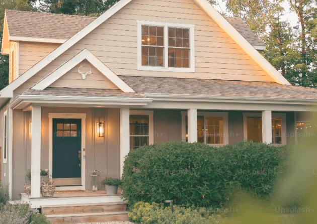 Maison familiale avec façade beige, porche accueillant et lumière allumée à l’entrée, entourée de verdure