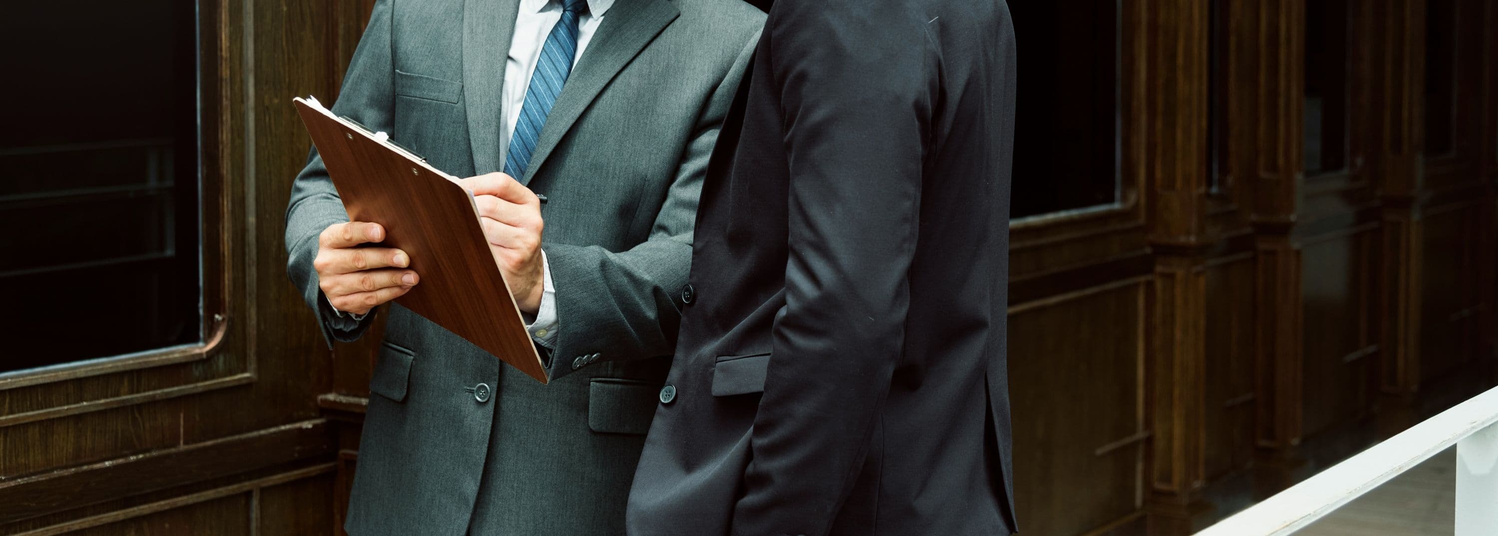 Deux hommes en costume discutant devant un bâtiment en bois, l’un tenant une planche avec des documents.